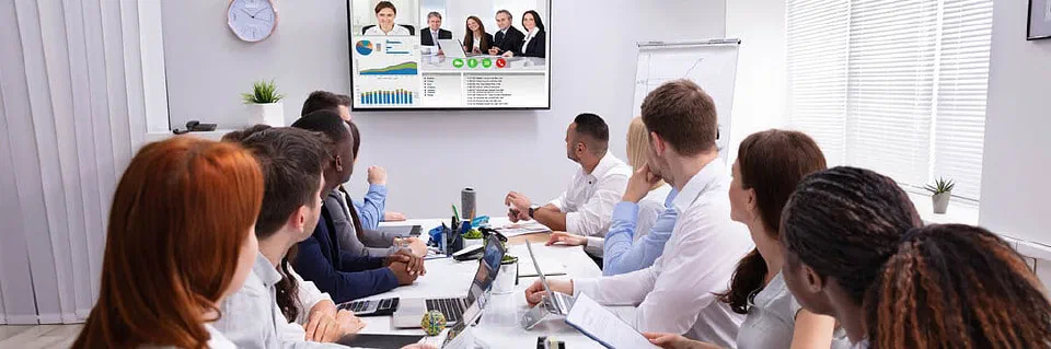 A group of people in a meeting room, conversing with a group of people online.